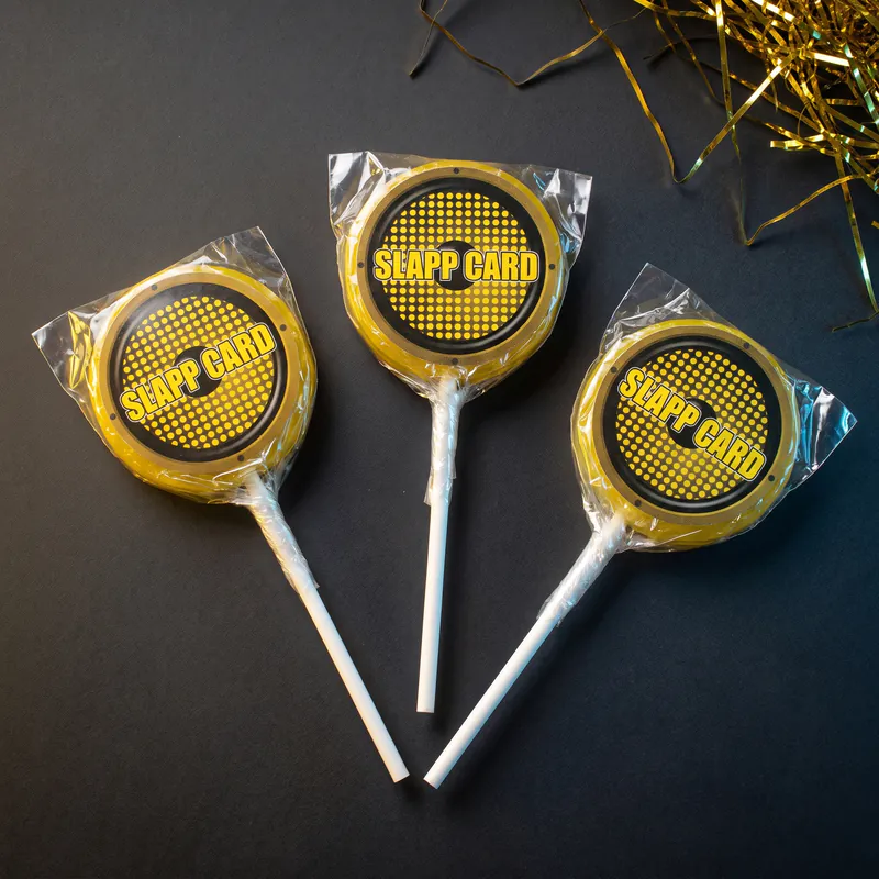 Three wrapped yellow suckers with the Slappcard logo on a dark surface, next to gold tinsel